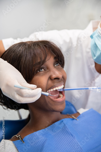 Young African American woman during cleaning procedure in clinic. Female dentist using hook and suction for cleaning or treating teeth. Oral hygiene and dental care concept