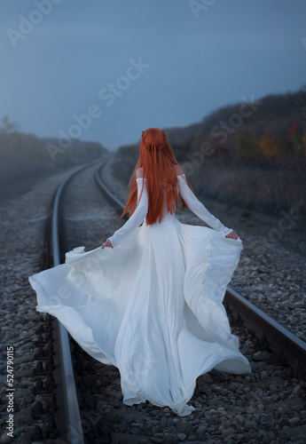 A woman with long red hair in a long white dress walks along the railway. Rear view. Vintage style. There is no face. A girl in retro style, going off into the distance by rail.