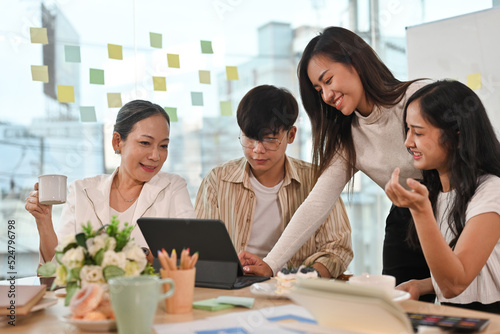 Attractive Asian senior businesswoman sitting at the office table with group of colleagues, Working on tablet computer, brainstorming, sharing ideas and solutions, negotiating on project.