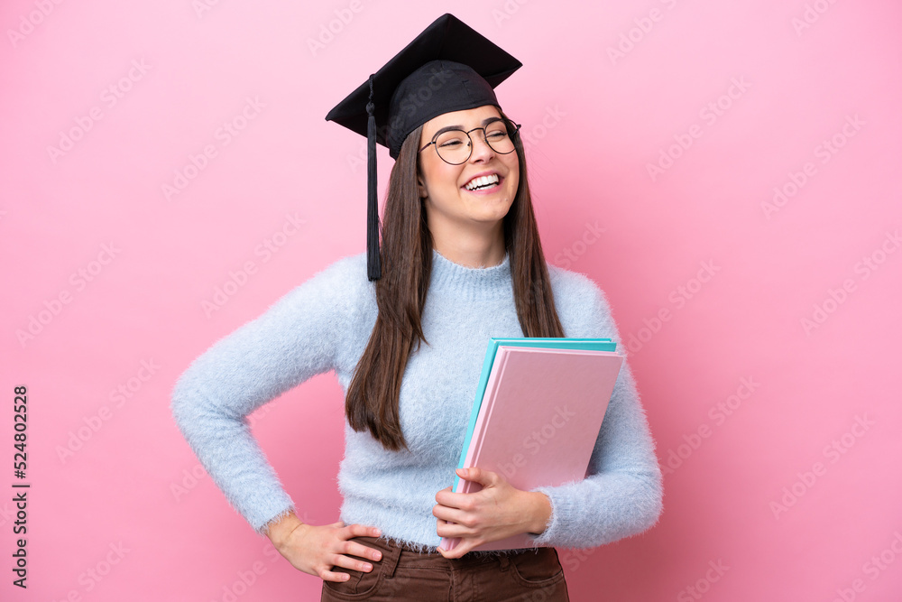 Young student Brazilian woman wearing graduated hat isolated on pink background posing with arms at hip and smiling