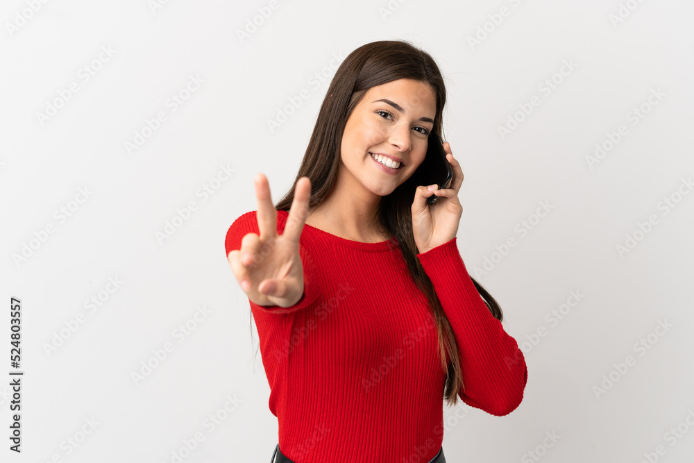 Teenager Brazilian girl using mobile phone over isolated white background smiling and showing victory sign Teenager Brazilian girl using mobile phone over isolated white background smiling and showing victory sign