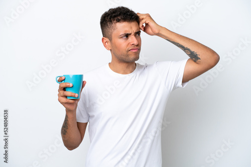 Young Brazilian man holding cup of coffee isolated on white background having doubts and with confuse face expression © luismolinero