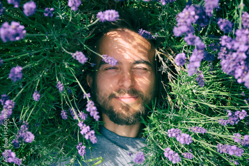 Portrait of young handsome bearded man lying among lavender flowers in blossom field. Happy smiling guy relax on the grass on sunny summer day. Freedom, love nature concept.