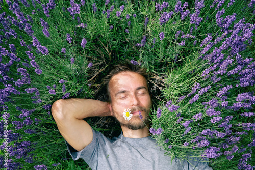 Portrait of young handsome bearded man lying among lavender flowers in blossom field. Happy smiling guy relax on the grass on sunny summer day. Self care mind body and soul.