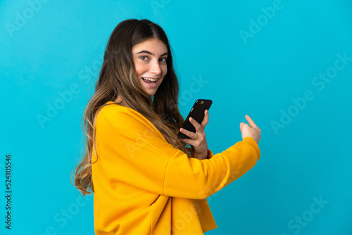 Young caucasian woman isolated on blue background using mobile phone and pointing back