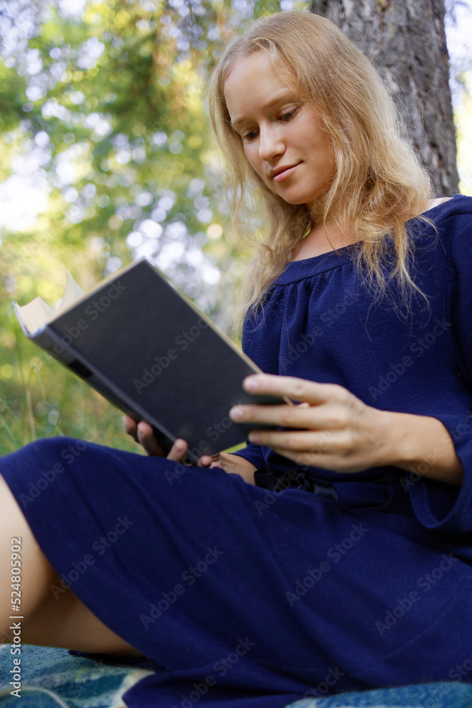 Obraz premium The girl reads a book in a park sitting under a tree. Shallow focus.