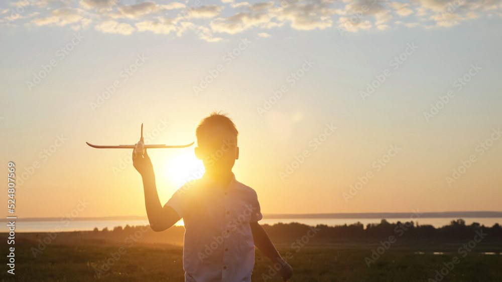 Happy boy runs on evening meadow playing with toy airplane at back sunset. Junior schoolboy silhouette enjoys playing with toys against blue sky