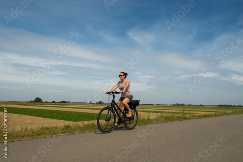 A young lady riding her bicycle on a cycle path through very typical Dutch countryside in the Netherlands on a clear sunny summer day. Example of uniform precision farming can be seen behind