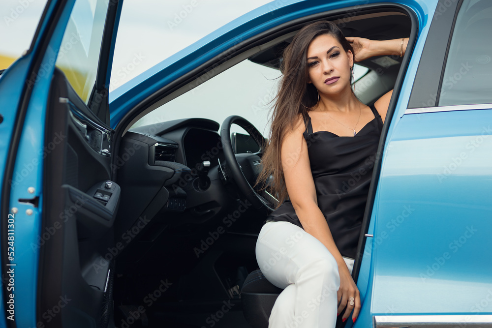 Successful woman driver demonstrates confident pose wearing elegant suit. Black-haired businesswoman sits in modern blue car at steering wheel