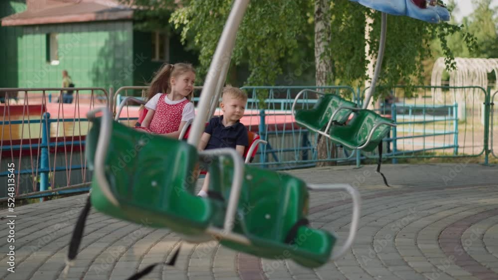 Little children ride marry-go-round attraction in summer park slow motion. Boy and girl have fun in amusement city park. Safe entertainment in town Little children ride marry-go-round attraction in summer park slow motion. Boy and girl have fun in amusement city park. Safe entertainment in town