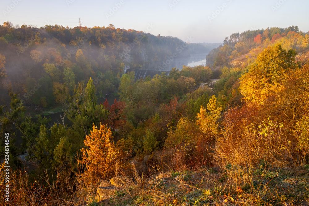 Autumn scenery with colorful trees on the rocky hillsides of Teteriv river banks. Yellow and red foliage illuminated with rising sun. Zhytomyr, Ukraine.
