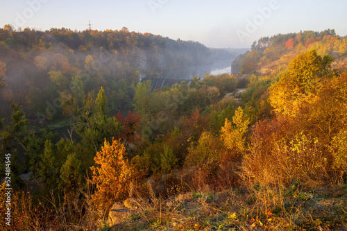 Autumn scenery with colorful trees on the rocky hillsides of Teteriv river banks. Yellow and red foliage illuminated with rising sun. Zhytomyr, Ukraine.