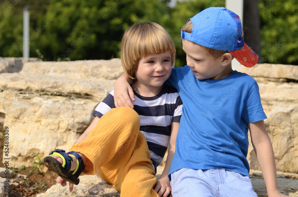 Two little boys, two friends, brothers are sitting on the street embracing. Concept: friendship, family, adventure, childhood, preschool education, kindergarten, amusement park, traveling with childre Two little boys, two friends, brothers are sitting on the street embracing. Concept: friendship, family, adventure, childhood, preschool education, kindergarten, amusement park, traveling with childre