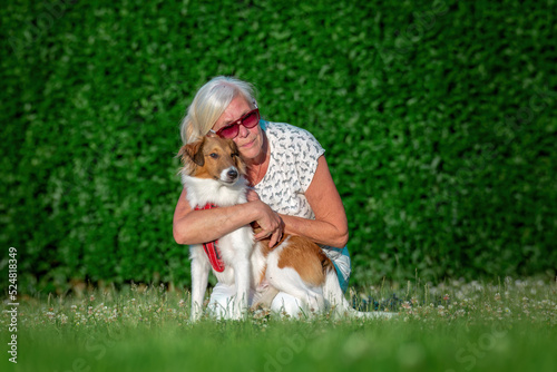  Older lady wearing sunglasses cuddling young sheltie puppy dog in the park behind a green background.