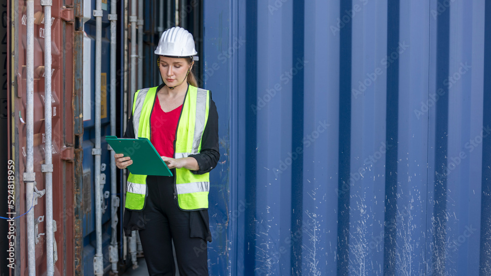 Young female inspector in safety vest checking stocks and goods in a logistic shipyard. Worker with helmet and reflective jacket reads file next to a cargo container Young female inspector in safety vest checking stocks and goods in a logistic shipyard. Worker with helmet and reflective jacket reads file next to a cargo container