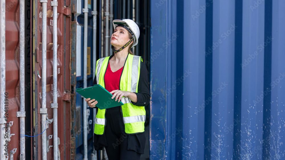 Young female inspector in safety vest checking stocks and goods in a logistic shipyard. Worker with helmet and  reflective jacket reads file next to a cargo container