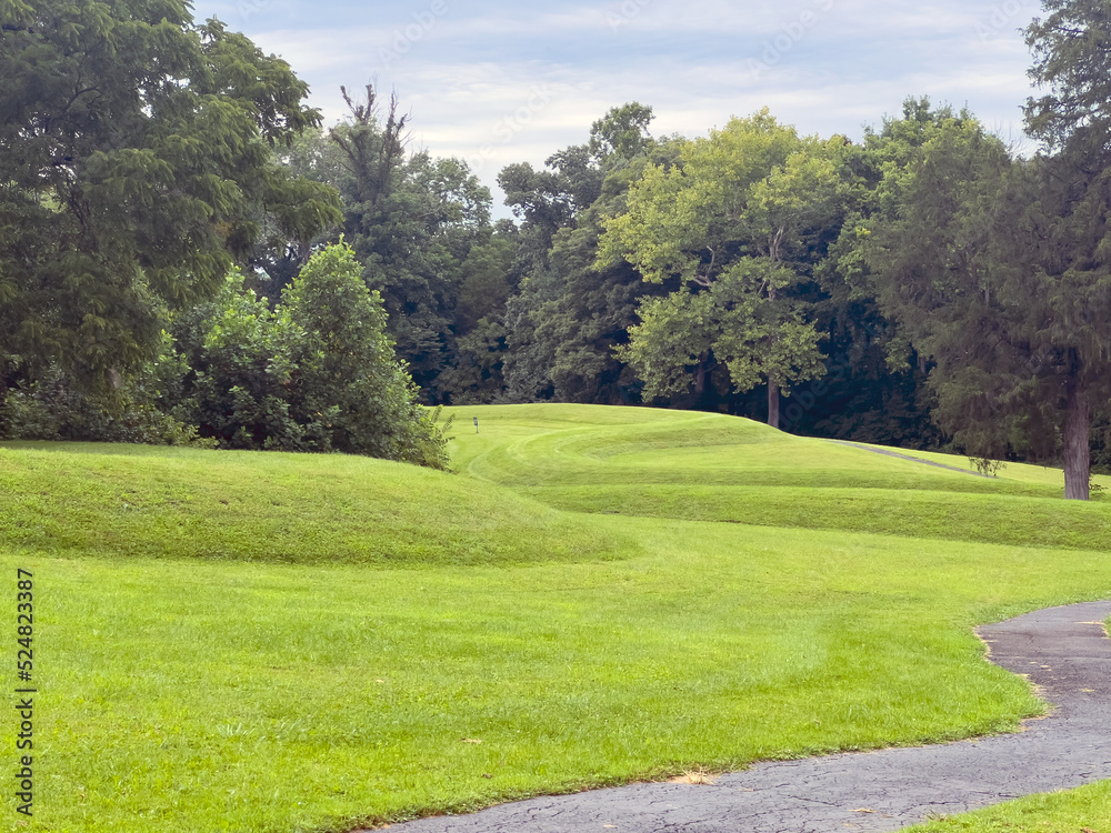 Walking path and curved earthworks prehistoric Great Serpent Mound. Body of snake effigy visible along landscape with woods and dramatic sky.