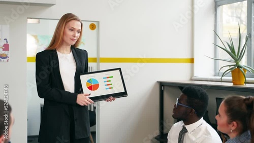 Business woman presents report, shows diagrams on tablet pc in conference room for multiethnic partners. Diverse international team listen CEO on board meeting in office. Corporate people discuss plan photo