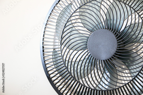 Close up of spinning black electric fan showing its rotating vanes and blades  on right of frame  against white wall background  necessary appliance for summer heat waves and rising temperatures