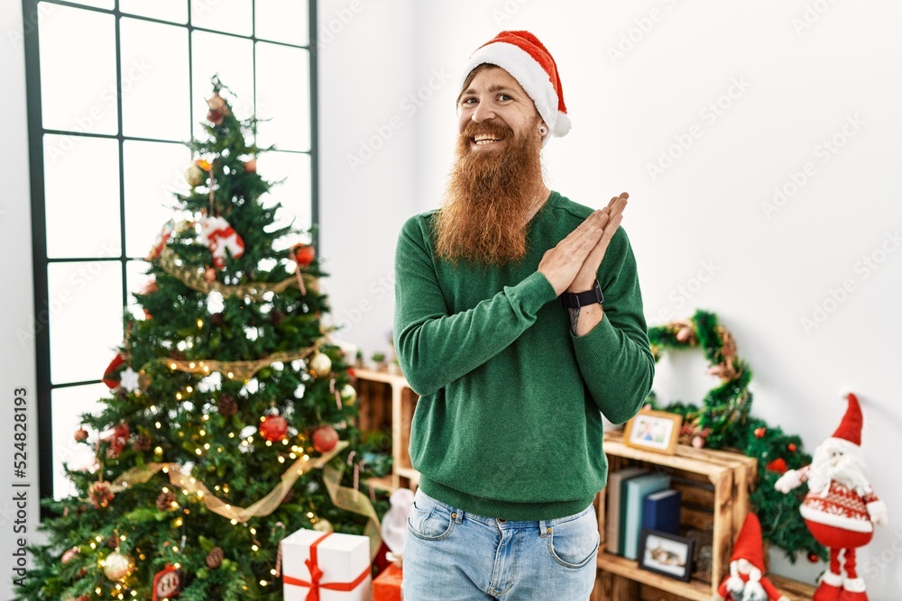 Redhead man with long beard wearing christmas hat by christmas tree clapping and applauding happy and joyful, smiling proud hands together Redhead man with long beard wearing christmas hat by christmas tree clapping and applauding happy and joyful, smiling proud hands together