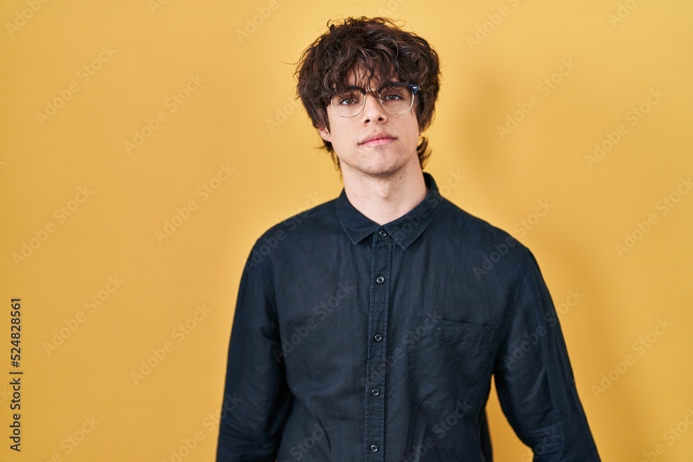Obraz premium Young man wearing glasses over yellow background relaxed with serious expression on face. simple and natural looking at the camera.