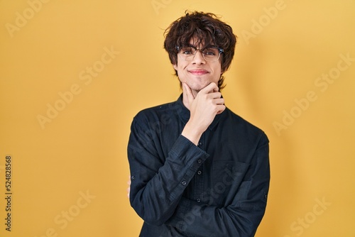 Young man wearing glasses over yellow background looking confident at the camera smiling with crossed arms and hand raised on chin. thinking positive. © Krakenimages.com