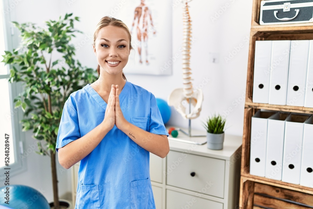 Young caucasian woman working at pain recovery clinic praying with hands together asking for forgiveness smiling confident. Young caucasian woman working at pain recovery clinic praying with hands together asking for forgiveness smiling confident.