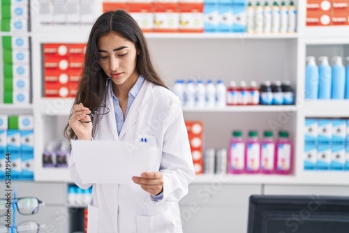 Young hispanic girl pharmacist reading prescription at pharmacy