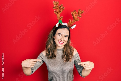 Print op canvas Young hispanic girl wearing deer christmas hat looking confident with smile on face, pointing oneself with fingers proud and happy