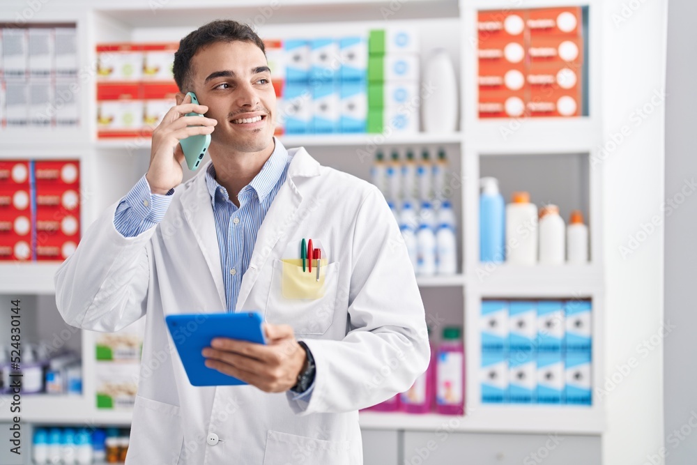 Young hispanic man pharmacist talking on smartphone using touchpad at pharmacy