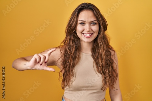 Young hispanic woman standing over yellow background smiling and confident gesturing with hand doing small size sign with fingers looking and the camera. measure concept.