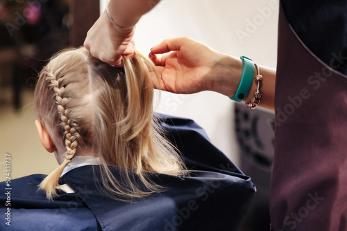Hair salon, hairdresser makes hairdo braids pigtail for young baby in barber shop. Barber woman make fashionable hairstyle for cute little blond girl child in modern barbershop. Copy space