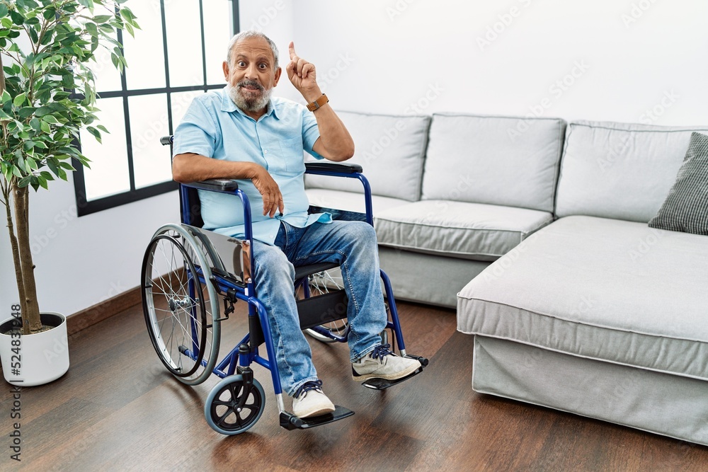 Handsome senior man sitting on wheelchair at the living room pointing finger up with successful idea. exited and happy. number one. Handsome senior man sitting on wheelchair at the living room pointing finger up with successful idea. exited and happy. number one.