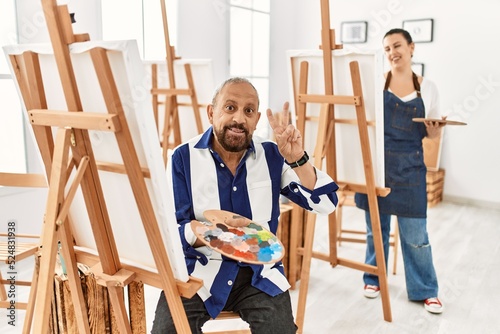 Senior artist man at art studio smiling looking to the camera showing fingers doing victory sign. number two.