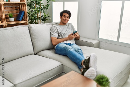 Young hispanic man using smartphone sitting on the sofa at home.