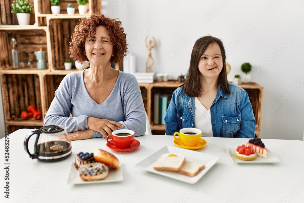 Family of mother and down syndrome daughter sitting at home eating breakfast winking looking at the camera with sexy expression, cheerful and happy face.