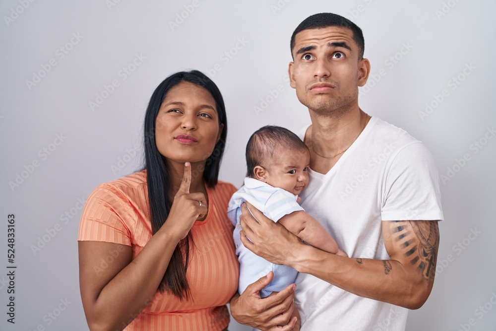 Young hispanic couple with baby standing together over isolated background thinking concentrated about doubt with finger on chin and looking up wondering