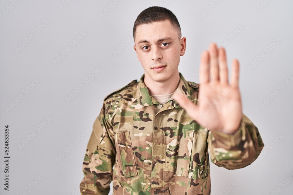 Young man wearing camouflage army uniform doing stop sing with palm of the hand. warning expression with negative and serious gesture on the face. Young man wearing camouflage army uniform doing stop sing with palm of the hand. warning expression with negative and serious gesture on the face.