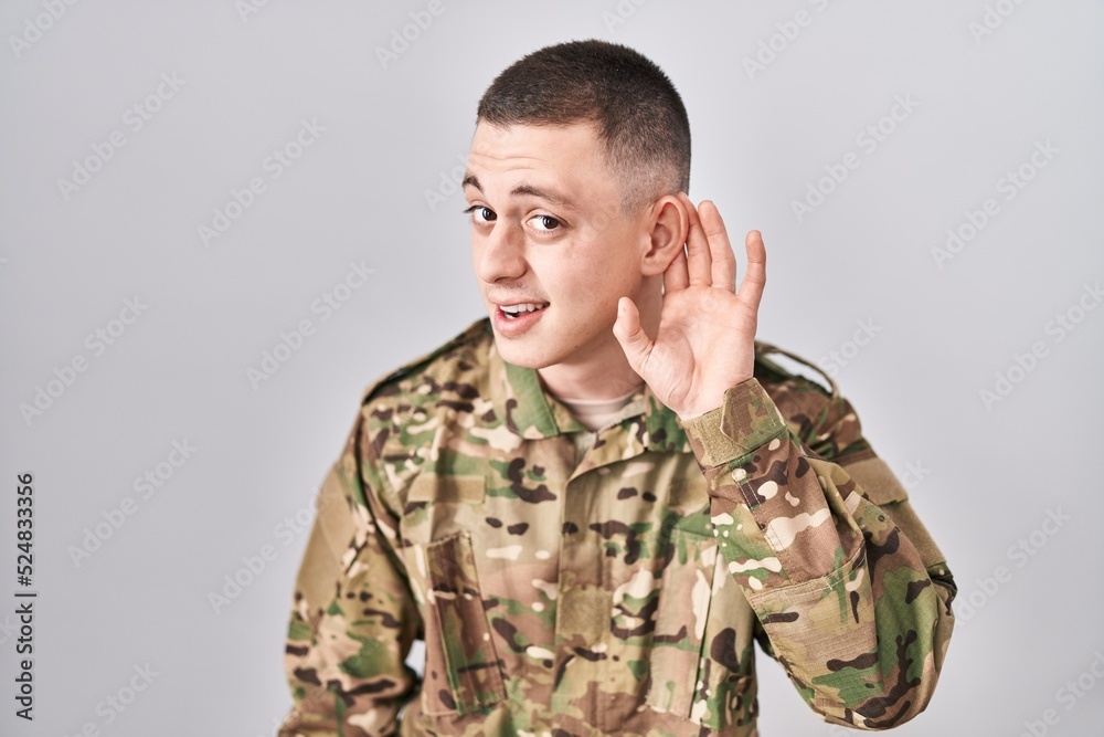 Young man wearing camouflage army uniform smiling with hand over ear listening an hearing to rumor or gossip. deafness concept.