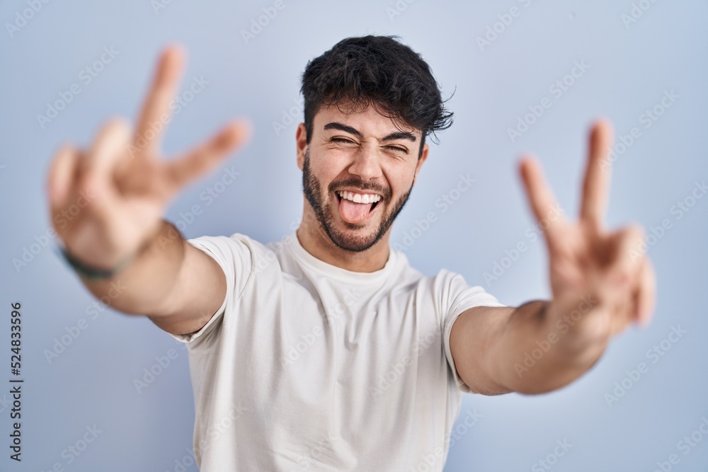 Hispanic man with beard standing over white background smiling with tongue out showing fingers of both hands doing victory sign. number two. Hispanic man with beard standing over white background smiling with tongue out showing fingers of both hands doing victory sign. number two.