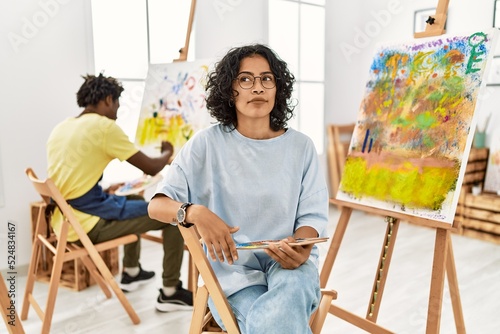 Young hispanic woman at art studio smiling looking to the side and staring away thinking.