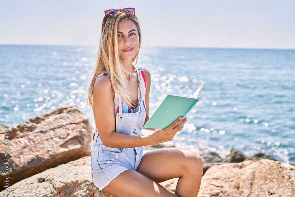 Young blonde girl reading book sitting on the rock at the beach.
