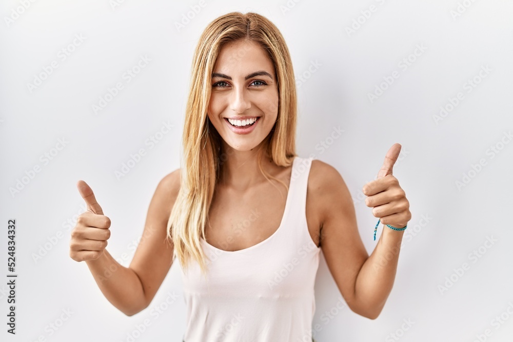 Blonde beautiful young woman standing over white isolated background success sign doing positive gesture with hand, thumbs up smiling and happy. cheerful expression and winner gesture. Blonde beautiful young woman standing over white isolated background success sign doing positive gesture with hand, thumbs up smiling and happy. cheerful expression and winner gesture.