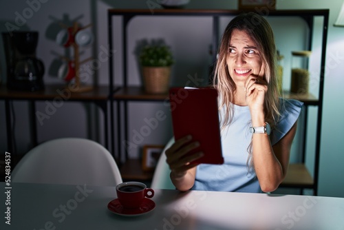 Young hispanic woman using touchpad sitting on the table at night thinking worried about a question, concerned and nervous with hand on chin © Krakenimages.com