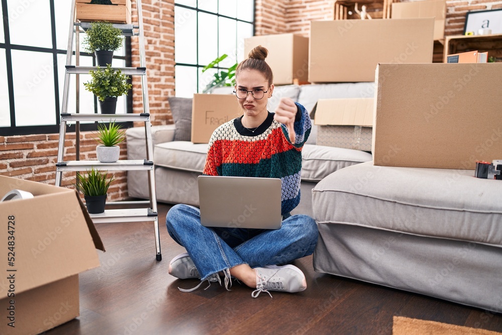 Young hispanic girl sitting on the floor at new home with laptop with angry face, negative sign showing dislike with thumbs down, rejection concept Young hispanic girl sitting on the floor at new home with laptop with angry face, negative sign showing dislike with thumbs down, rejection concept