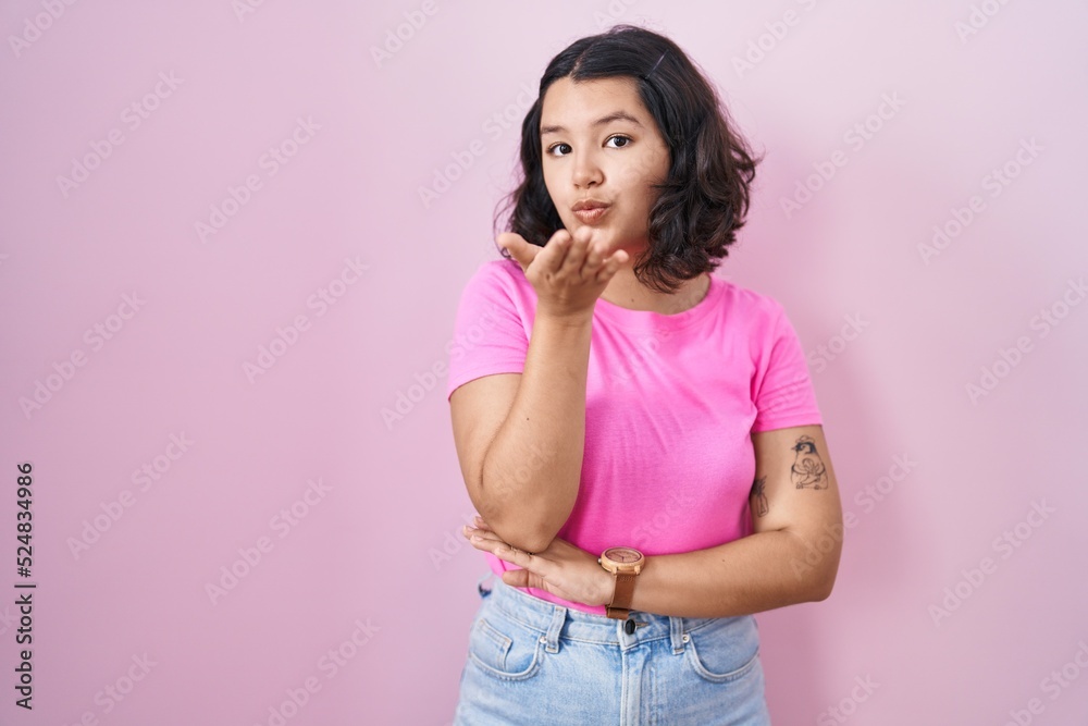 Young hispanic woman standing over pink background looking at the camera blowing a kiss with hand on air being lovely and sexy. love expression. Young hispanic woman standing over pink background looking at the camera blowing a kiss with hand on air being lovely and sexy. love expression.
