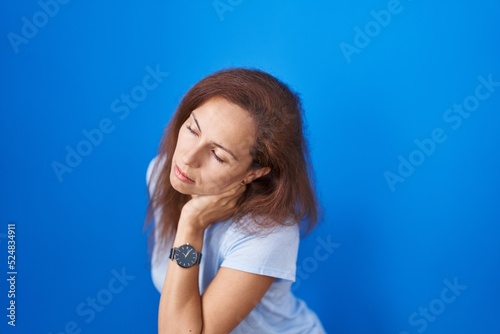 Brunette woman standing over blue background suffering of backache, touching back with hand, muscular pain