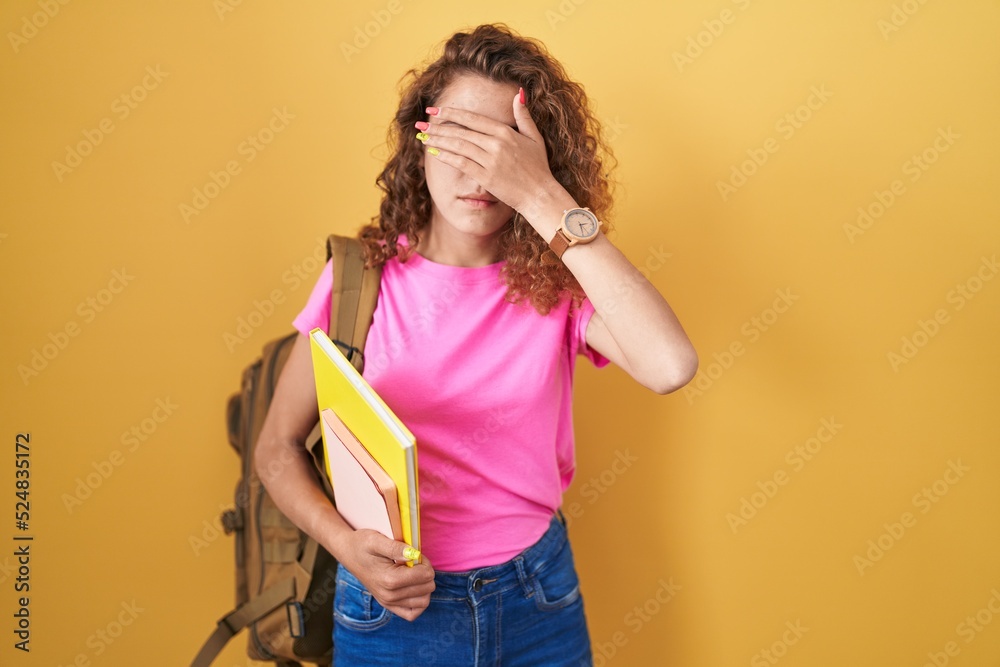 Young caucasian woman wearing student backpack and holding books covering eyes with hand, looking serious and sad. sightless, hiding and rejection concept Young caucasian woman wearing student backpack and holding books covering eyes with hand, looking serious and sad. sightless, hiding and rejection concept