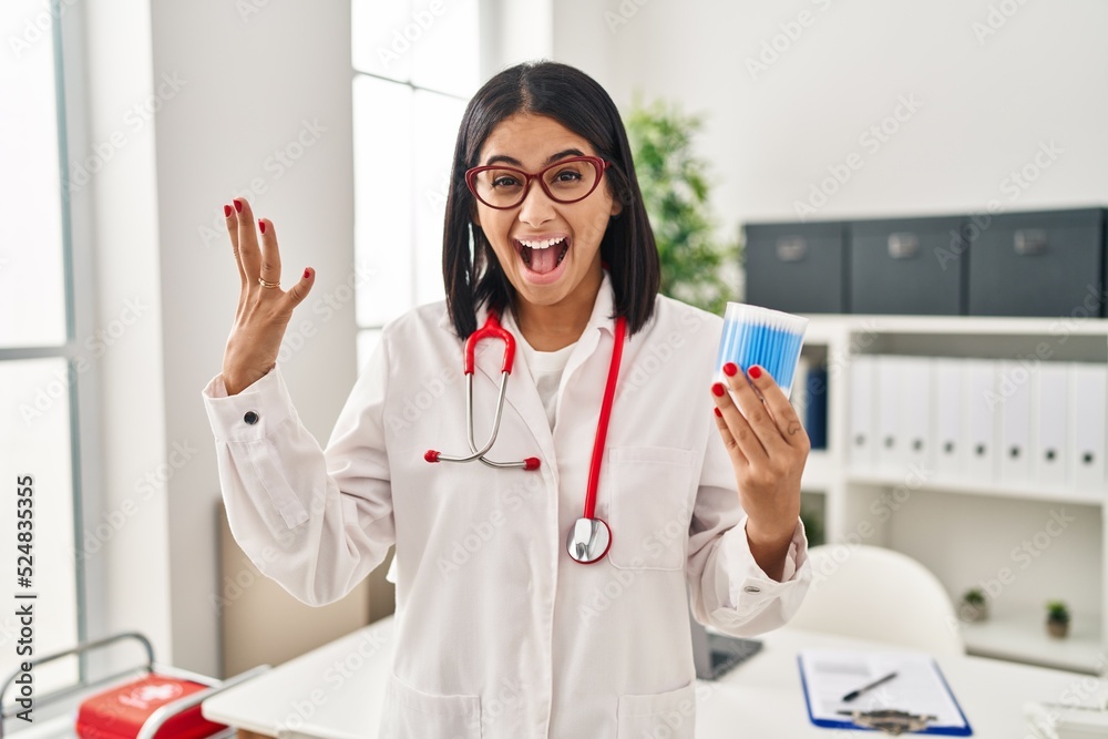 Young hispanic doctor woman holding cotton buds celebrating victory with happy smile and winner expression with raised hands