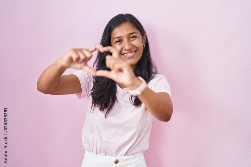 Young hispanic woman standing over pink background smiling in love doing heart symbol shape with hands. romantic concept.